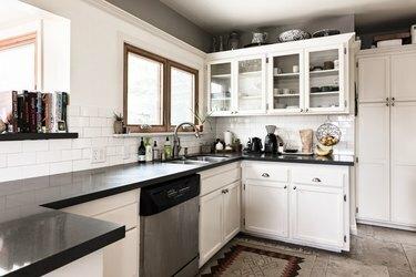 virtuvė su baltomis virtuvės spintelėmis ir stikliniais įdėklais, tamsiais stalviršiais ir metro plytelių backsplash
