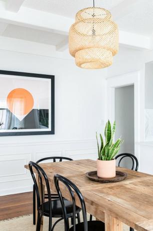 ideia de decoração de parede de sala de jantar com mesa de jantar de madeira, planta de cobra, luz pendente de vime e arte