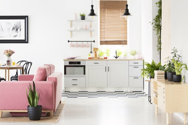 Foto real do interior da cozinha de espaço aberto com piso quadriculado, janela com persianas de madeira, sofá de veludo rosa e muitas plantas frescas