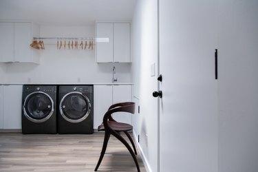 Модернистичен дървен стол в минималистично перално помещение с бели шкафове и черна пералня и сушилня.