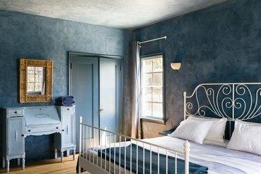 Spavaća soba s plavim zidovima, bijelim spiralnim okvirom kreveta, plavom posteljinom, vintage plavim stolom, retrovizorom od zlatnog zlata, dvokrilnim vratima i prozorskim zavjesama.
