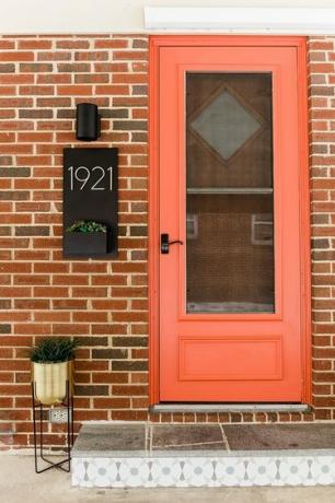 Forreste indgang til et hus med en orange dør, sort sconce lys, guld plantestand, mursten udvendigt