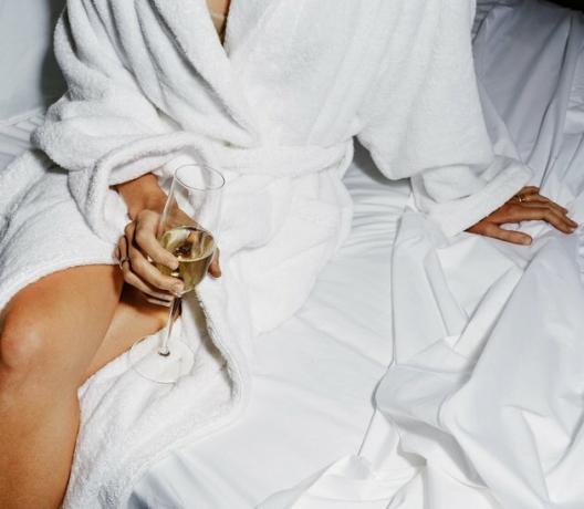 Een vrouw in een badjas op een wit bed met een glas champagne in haar handen.