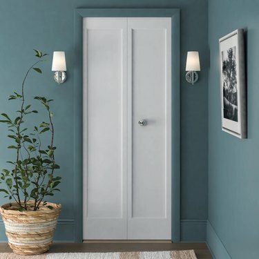 Hvide tofoldede døre i et blågrønt rum; hvide lampetter er på hver side af døren og en stor plante er til venstre for skabet