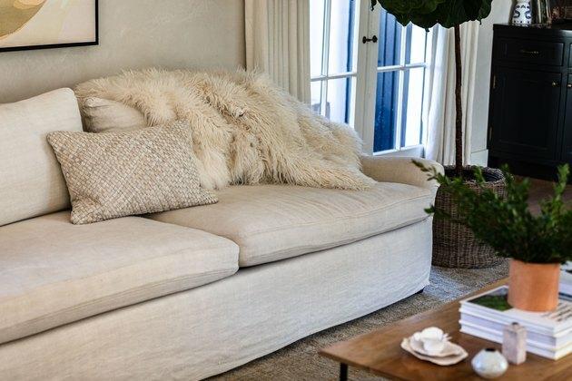 Balta svetainės sofa su derančia pagalve ir balta kailinė antklodė prie kavos stalo