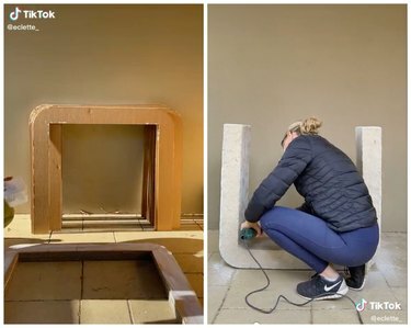 Aan de linkerkant is een stapel karton in de vorm van een tafel die tegen een muur leunt. Rechts staat een vrouw in een marineblauw jasje een tafel aan het schuren.