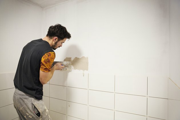 Un constructor, tiler care plasează plăci ceramice albe pe un perete într-o baie.