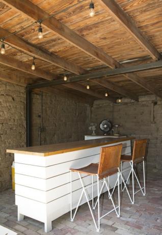 Bar contemporâneo com balcão de madeira, luzes de corda, teto com viga de madeira e ventilador industrial