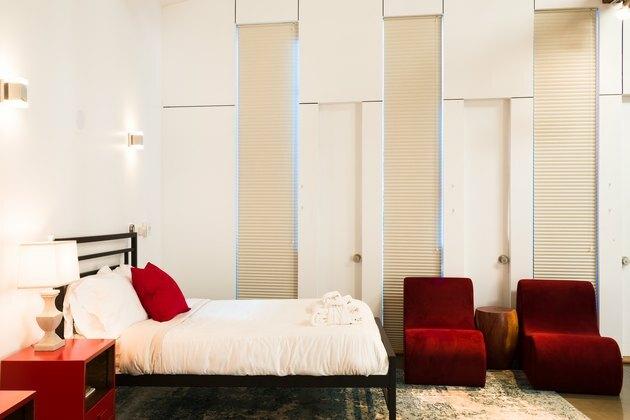 Quarto de hotel com cama queen-size e cabeceira preta, mesa lateral vermelha e poltronas sem braços