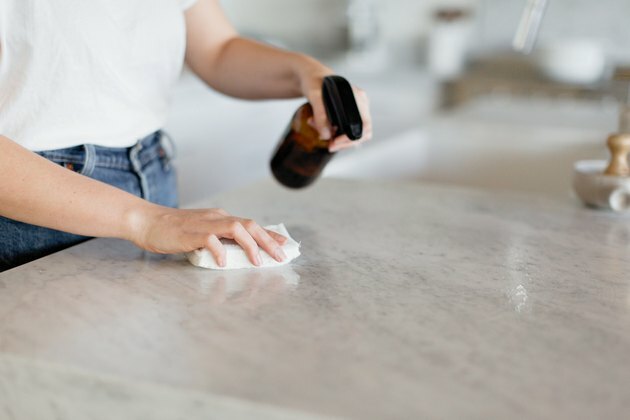 Foto do meio e das mãos de uma mulher em uma camiseta branca e jeans. Ela está limpando o balcão da cozinha com um spray de vidro marrom em uma das mãos e um pano branco na outra.