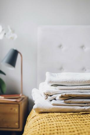 Lençóis brancos dobrados em uma cama ao lado de uma mesa de cabeceira de madeira com abajur