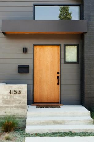 O casă gri închis, cu accente negre și o ușă de intrare din lemn deschis. Ferestre variate si trepte din fata din beton.