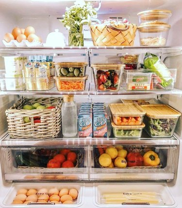 Alimentos e bebidas organizados em lixeiras e cestos transparentes dentro de uma geladeira.