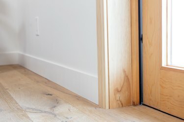Drevená podlaha pri dverách. Spodná časť drevenej zárubne vedľa bielej steny s bielym lemovaním.
