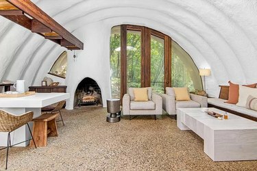 Гостиная с белыми сводчатыми потолками и коричневыми акцентами. В углу камин и частично видна белая кухня.