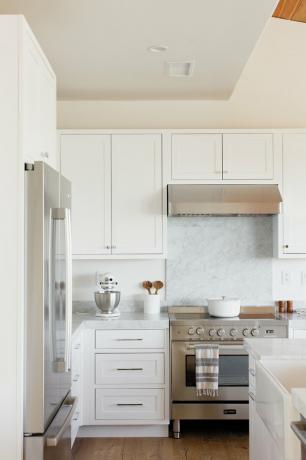 Cozinha com gama de inox, armários brancos, recuperador de calor e bancada em mármore. Na bancada, há uma batedeira KitchenAid branca com uma tigela de aço inoxidável e um porta-utensílios branco com pinças de madeira para salada. No fogão, há um forno holandês branco. À esquerda, há uma geladeira de aço inoxidável. À direita, há uma ilha de cozinha branca com pia.