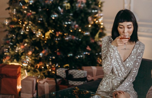 mulher no sofá com um vestido brilhante tomando um coquetel com árvores de natal e presentes no fundo