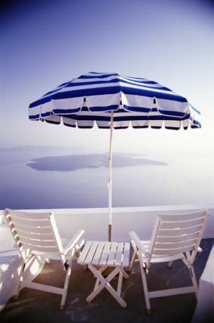 Bakre utsikt over trestoler og et bord med paraply på en balkong med utsikt over havet og de greske øyer.