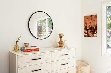 خزانة بيضاء مع مزهرية بيج ونحت طائر مع مرآة مستديرة وفن حائط معاصر