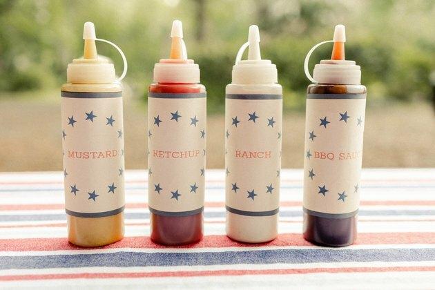 Keturi dešrainių barų prieskonių buteliai su atspausdintomis garstyčių, kečupo, rančos ir BBQ padažo etiketėmis
