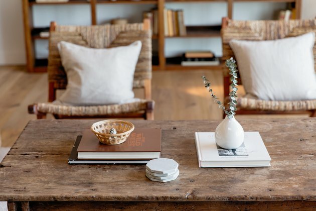 Duas pilhas de livros sobre uma mesa de centro rústica. Em uma delas, um pequeno vaso branco com eucalipto. Do outro, uma pequena cesta de tecido. Há também uma pilha de bases para copos de cerâmica cinza. Atrás da mesa de centro, duas cadeiras com almofadas em tons de cinza claro.