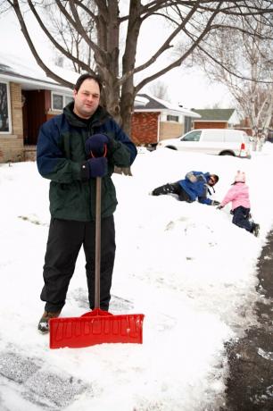 Tatăl pozând cu lopata de zăpadă în timp ce copiii se joacă