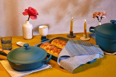 Il set Shabbat di Our Place comprende una teglia verde acqua con challah coperto da un panno, due candelieri, una stuoia da forno, un vaso con un fiore rosa tutto su un tavolo di legno