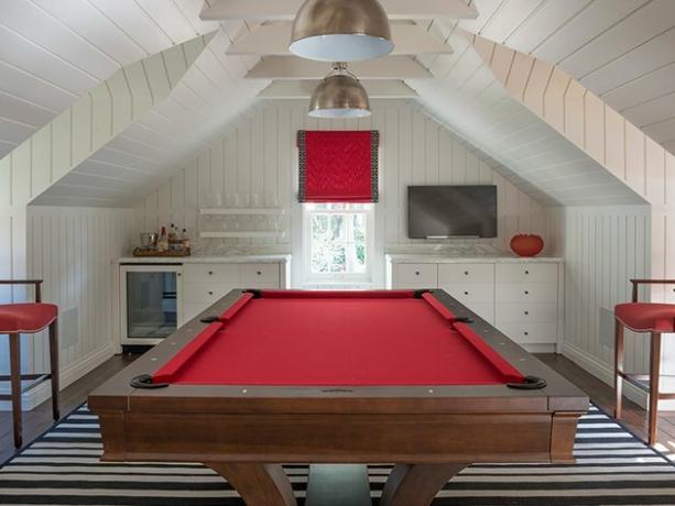 zolder speelkamer met rode pooltafel, rode krukken, rode raamschaduw, witte scheepswanden en plafond, metalen hanglampen, zwart-wit gestreepte tapijten, flatscreen, ingebouwde kasten.