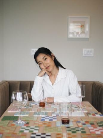 Grace Kim, uma mulher asiático-americana, vestindo uma camisa branca de botão, sentada em uma mesa coberta com seus porta-copos feitos à mão