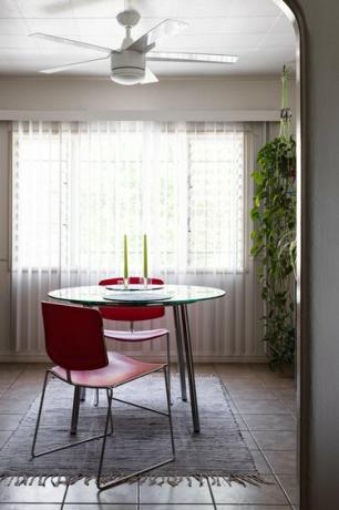 To røde stoler ved et rundt spisebord med to lysestaker. Rene gardiner, hengende anlegg og takvifte.