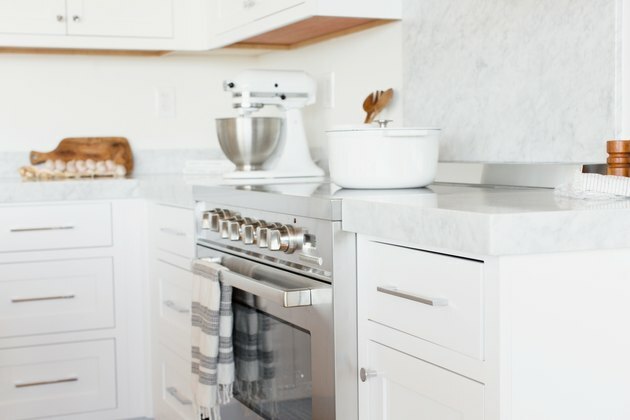 Balcão da cozinha com gavetas brancas e bancada em mármore branco. No meio do balcão, uma gama de aço inoxidável. Um pano de prato listrado de branco e cinza está pendurado na maçaneta da porta do forno. Um forno holandês branco está sobre o fogão, e há uma batedeira KitchenAid branca.