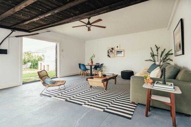 Idéias para pequenas garagens em uma sala de estar no estilo boêmio da Califórnia: piso de cimento, teto de madeira, ventilador de teto, tapete preto e branco, cadeira de vime, planta, mesa de centro e sofá. Uma garagem renovada com área de estar, piso de cimento e teto de madeira.
