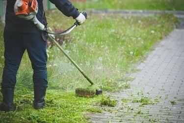 Työntekijä tummissa suojavaatteissa kaasuleikkuri käsissään, leikkaa ruohoa talon edessä. Mies leikkaa ruohoa voikukkien kanssa sateisena kevätpäivänä. Trimmeri miehen käsissä.