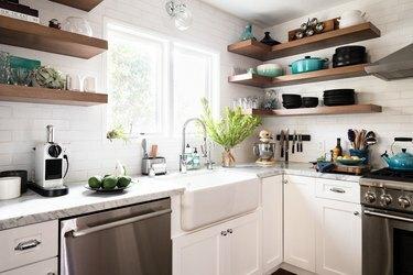 Uma área de cozinha com armários brancos, lava-louças de aço inoxidável, forno e prateleiras abertas de madeira empilhadas com utensílios pretos, brancos e azuis.