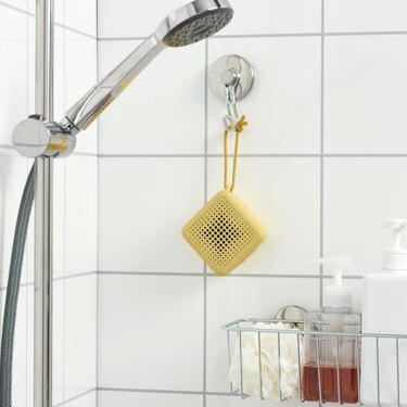 רמקול צהוב של איקאה VAPPEBY תלוי במקלחת אריחים לבנים מעל מדף של מוצרי טיפוח למקלחת.