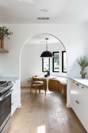 Vista della sala da pranzo con panca curva, tavolino rotondo, sedie, lampada a sospensione e alte finestre attraverso l'arco dallo spazio cucina con pareti bianche