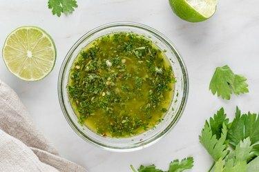 La salsa chimichurri in una ciotola di vetro trasparente, ancora contornata da due metà di lime e coriandolo.