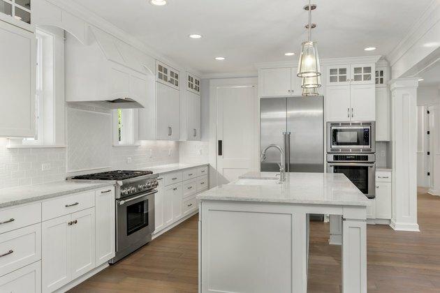 nuostabi balta virtuvė naujuose prabangiuose namuose su sala, pakabinamais žibintais ir kietmedžio grindimis. Pasižymi nerūdijančio plieno prietaisais ir sodybos stiliaus kriaukle