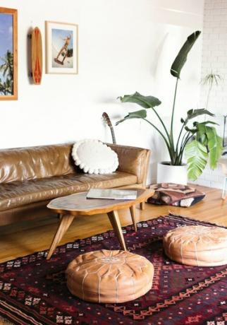 Pouf in pelle con ricamo bianco bianco, su tappeto tradizionale. Decorazioni per skateboard, tavolo in legno, divano in pelle e piante.