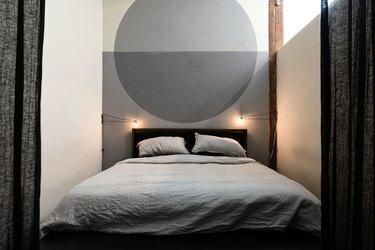 Entrada con cortinas negras que conduce a un dormitorio industrial minimalista con un mural circular gris, ropa de cama gris y apliques de alambre industriales.