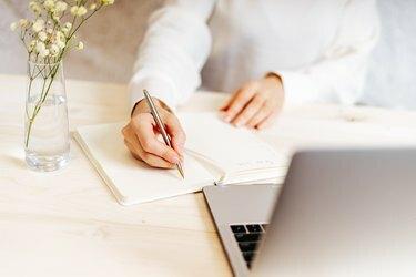امرأة تكتب في دفتر ملاحظات على مكتب به كمبيوتر محمول