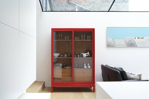 غرفة بيضاء حديثة مع كوة كبيرة ، وطلاء ، وأرضيات خشبية فاتحة وخزانة عرض ملونة بلون فيرميون بأبواب زجاجية.