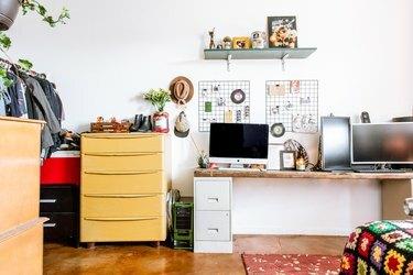 Computadores em uma mesa com mesa de madeira e arquivos brancos. Armário amarelo, ao lado. Quadros de arame, chapéus e plantas como decoração.