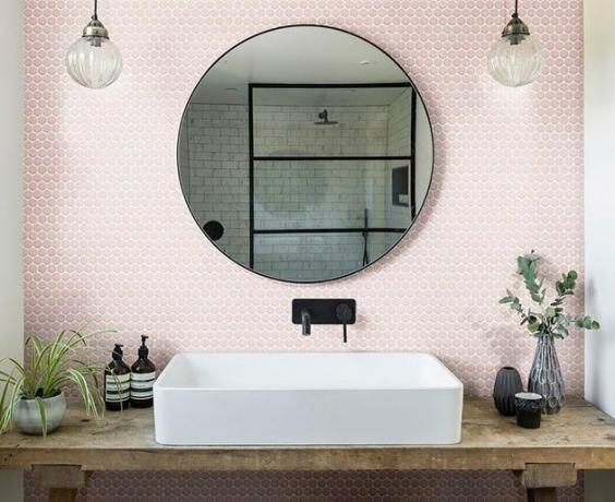 Banheiro rústico moderno com pia branca, backsplash de hectaganol rosa, torneira de banheiro fixada na parede preta, lâmpadas globo, penteadeira de madeira.