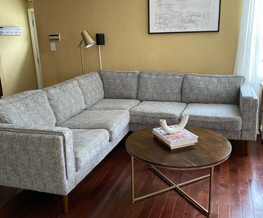 Um corte cinza em uma sala amarela; uma mesa de centro redonda marrom com livros e um vaso decorativo em cima