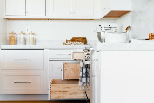 Na cozinha, gavetas brancas com bancada de mármore e armários brancos acima. Três das gavetas estão abertas em vários graus. Na bancada, há uma batedeira KitchenAid branca, três recipientes de armazenamento de vidro e uma panela branca.