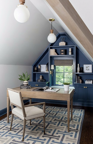 Apartamento no sótão com parede azul-escura, prateleiras embutidas, mesa, cadeira, tapete estampado, luminárias pendentes em forma de globo branco.