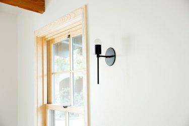 Uma janela com moldura de madeira clara contra uma parede branca, perto do canto da sala. Um bolinho de metal preto está ao lado da janela.