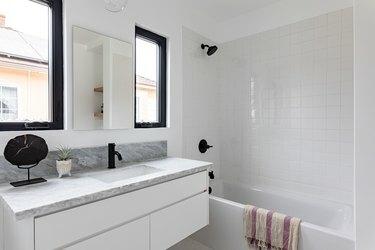 Valkoinen kaakeloitu kylpyhuone, jossa kylpyamme, musta suihkupää ja hana, mustat kehystetyt ikkunat ja marmorinen työtaso