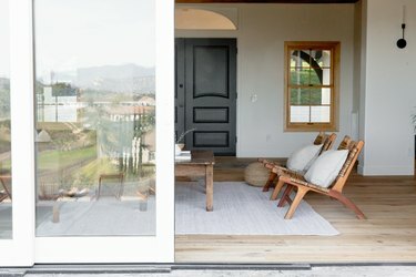 Uși glisante din sticlă cu cadru alb care duc la un living unde sunt vizibile două scaune din lemn de tec cu perne decorative și o măsuță de cafea rustică.